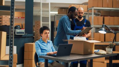 Diverse startup partners doing quality control before shipping goods order, packing merchandise in cardboard boxes. Workers team preparing delivery and shipment in warehouse storage space.