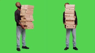 Genç insan kameraya büyük bir pizza yığını teslim ediyor, fast food siparişiyle birlikte bir sürü karton kutu taşıyor. Tam vücut yeşil ekran arkaplanı üzerinde poz veren paketleri olan yetişkin bir erkek.