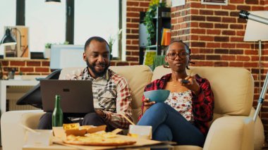 Happy modern people watching tv together at home, man browsing internet on laptop and woman eating bowl of snacks. Young couple enjoying comedy film with alcohol bottles. Tripod shot.