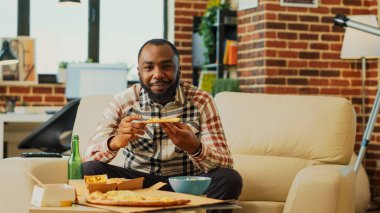 African american man eating slice of pizza at home, binge watching action tv series in living room. Modern happy guy enjoying fast food delivery and having fun with movie. Tripod shot.