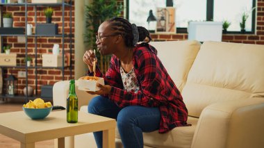 Cheerful girl serving noodles in delivery box, using chopsticks to eat asian food and watch action film on tv. Woman enjoying dinner from takeaway restaurant, having fun with television.