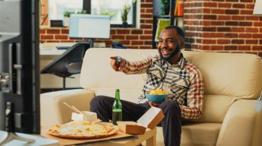 Young adult drinking bottle of beer and eating chips, having drinks and snacks at television. Cheerful relaxed man binge watching tv show in living room, enjoying fast food delivery.