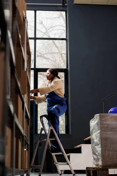 非裔美国妇女仓库工人站在梯子上 从高高的架子上拿起纸板箱 邮政局职员在分发库房的储藏室里寻找顾客邮包 — 图库照片