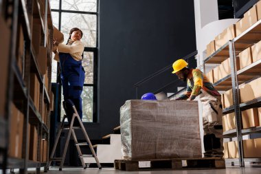 Depo müdürü merdivende duruyor ve müşteri siparişi için karton kutu alıyor. Postane çalışanı elinde karton kutuyla depodaki malları kontrol eden bir adam.