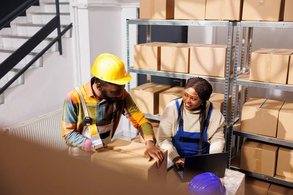 物流センターの労働者は ラップトップ上の商品在庫をチェックし 出荷のための貨物を梱包する 段ボール箱でいっぱいの倉庫で働くアフリカ系アメリカ人の物流管理者 — ストック写真