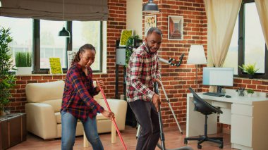 People of african ethnicity being funny and cleaning in living room, using mop to wash dirt and vacuum to clean floors. Young partners dancing and doing spring cleaning. Handheld shot.