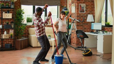 Happy man and woman doing house chores and dancing, listening to music for spring cleaning work. Cheerful couple sweeping floors and using vacuum, singing together in living room.