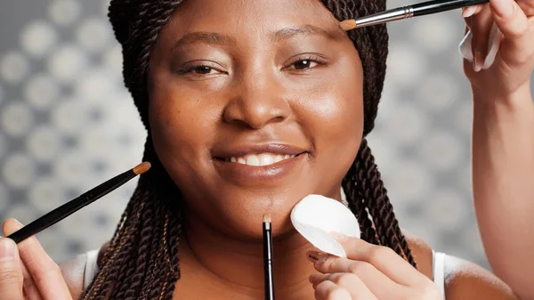 非洲裔美国妇女从化妆师团队 人们使用刷子和棉垫创造美丽的发光外观工作室化妆 选美模特儿为广告宣传作模特 靠近点 — 图库照片