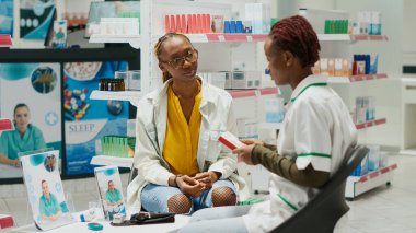 Female pharmacist showing box of pills to young client, giving pharmaceutical products in drugstore. Medic and woman talking about supplements and drugs in pharmacy shop. Tripod shot.