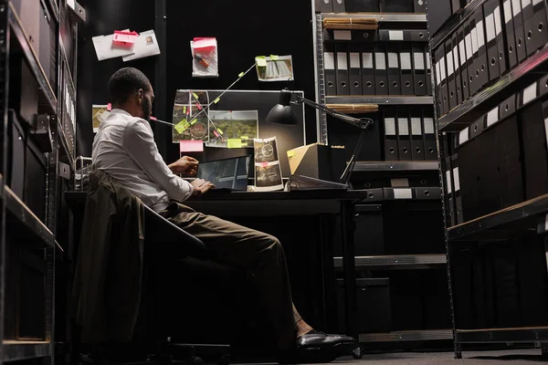 犯罪現場の監視写真をノートパソコンの画面で調べてる刑事証拠ボードの近くの机に座ってる アフリカ系アメリカ人の研究者は夜にコンピュータ上の写真を分析 — ストック写真