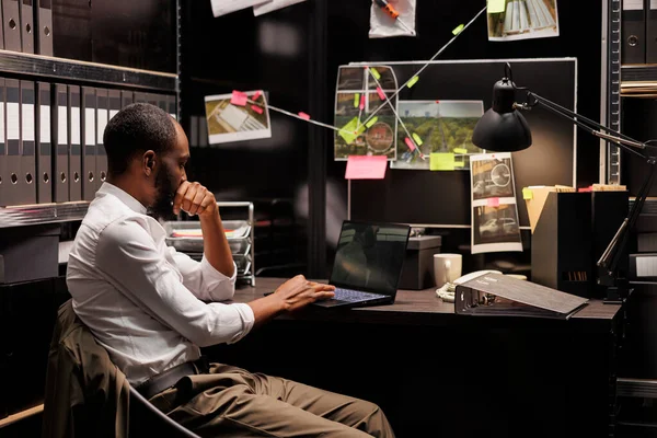 アフリカ系アメリカ人の私立探偵がノートパソコンで監視写真を分析して容疑者をスパイしてる 犯罪を解決するためにビデオ記録を見てオフィスのテーブルに座っている刑事 — ストック写真