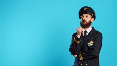 Male aviator in uniform thinking about solution, brainstorming ideas to make decision over blue backdrop. Young plane captain acting pensive and thoughtful, uncertain airliner.