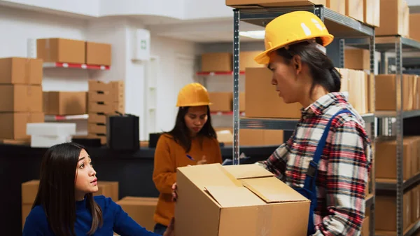Asian guy organizing boxes with supplies on shelves, doing quality control with female entrepreneur in warehouse. Employee putting packages on racks, preparing for order management.
