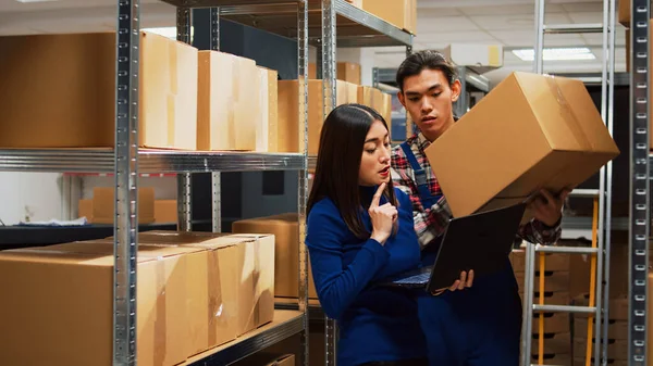 Team of people checking stock supplies on laptop, working with merchandise boxes in warehouse space. Man and woman doing teamwork for inventory and products logistics in storage room.
