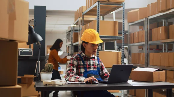 Asian person planning order management in warehouse, using laptop to do stock inventory. Male employee working with products in storage room, supply chain and distribution boxes.