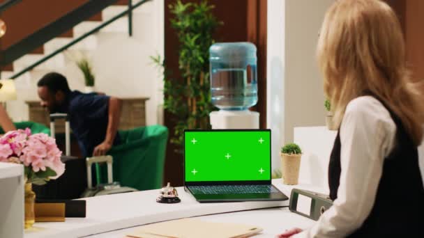 フロントのノートパソコンの緑のテンプレートを使用して フロントでお客様を歓迎ホテルの労働者 従業員はロビーで人々と話をし リゾートで孤立したモックアップディスプレイで働いています ティポッドショット — ストック動画