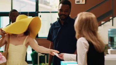 Gezginler lobideki resepsiyon görevlisine oda kartı verip tropikal deniz kıyısındaki tatil beldesinden ayrılıyorlar. Genç çift resepsiyonistle çıkış prosedürünü uyguluyor, vedalaşıyorlar. El kamerasıyla..