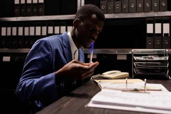 アフリカ系アメリカ人のマネージャーは 管理ファイルをチェックし 企業リポジトリの管理研究で夜遅くに働いています 貯蔵室の会計報告書を分析するビジネスマン — ストック写真