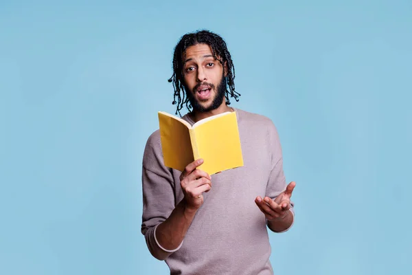 Arab man holding softcover book and explaining novel plot while looking at camera. Young person reading open paperback textbook with blank yellow cover and talking portrait