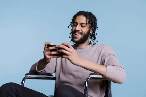 快乐的男人坐在轮椅上用智能手机玩手机游戏 使用具有愉快面部表情的电话应用程序对身体残疾的年轻人进行微笑 — 图库照片