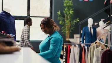 Yeni moda koleksiyonuna bakan Afrikalı Amerikalı müşteriler modern butikte şık kıyafetler alıyorlar. Moda ürünleri ile rafları kontrol eden şık bir istemci, mağazadaki kumaşı analiz ediyor