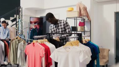 Afro-Amerikalı adam yeni moda koleksiyonu ile rafları kontrol ediyor, giyim mağazasında malzeme analizi yapıyor. Alışveriş hastası modern butikte şık kıyafetler satın alıyor.