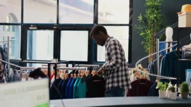 Afro-amerikan müşteriler modaya uygun gömlekli askılara bakıyorlar, giysileri almadan önce analiz ediyorlar. Alışveriş manyağı adam modern butikte şık ürünler ve aksesuarlar alıyor.