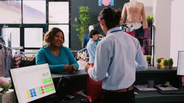 在现代精品店的柜台上 非洲裔美国人把信用卡放在顾客终端上支付时尚服装的费用 而妇女购物者则在服装店购买时髦商品 — 图库视频影像