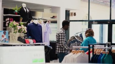 Afrikalı Amerikalı çift resmi kıyafet alışverişi yapıyor, modern butikten almadan önce modaya uygun kıyafet kumaşlarını kontrol ediyor. Giyim mağazasında yeni moda koleksiyonu olan askılara bakan şık insanlar.