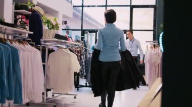 Neşeli bir çalışan şık kıyafetler ayarlıyor, modern bir butikte çalışıyor. Kafkasyalı yönetici, alışveriş merkezindeki raflara şık ürünlerle askı asıyor. Moda konsepti