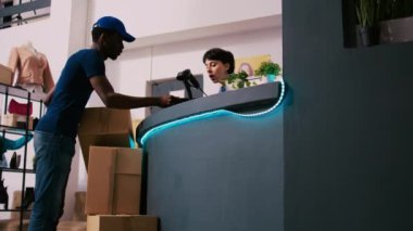 Afro-Amerikan kurye, nakliye raporunu işçiye gösteriyor, giysi mağazasında online sipariş detaylarını tartışıyor. Yönetici kasada duruyor, paketleri modern butikte nakliyat için hazırlıyor.