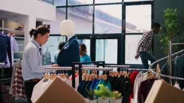 Asyalı yönetici mavi takım elbise düğmelerini kapatıyor, modern butikte şık kıyafetler ve şık aksesuarlar düzenliyor. Giyim mağazasında çalışan neşeli bir çalışan. Moda konsepti