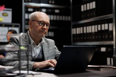 Üst düzey özel dedektif dizüstü bilgisayardaki gizli kriminoloji kanıt dosyalarını kontrol ediyor. Dolap raflarındaki klasörlerle çevrili ajans dosya depolama ofisindeki araştırmacı