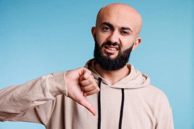 Arap adam olumsuz duygularla kameraya bakarken onaylamadığını göstermek için baş parmak hareketi yapıyor. Stüdyo portresindeki anlaşmazlıkları ifade eden genç bir insan.