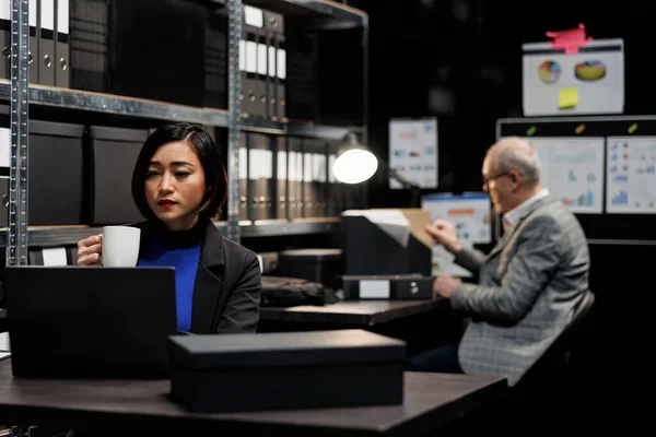 Accounting employees in consultancy bureaucratic workplace filled with business folders. Asian woman enjoying cup of coffee and senior coworker working in administrative file room office