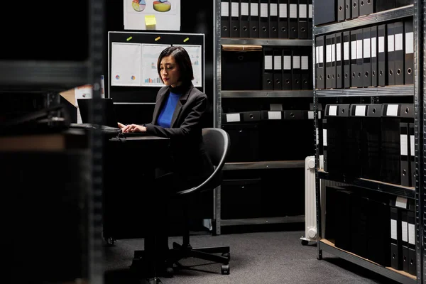 亚洲私家侦探检查笔记本电脑上的机密犯罪学证据文档 代理档案橱柜办公室的调查员被书架上的文件夹包围着 — 图库照片