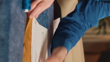Dikey video işyeri sahibi kıyafet yapıyor, özel yapım eşyalar yaratmak için terzilik atölyesindeki çizimlere bakıyor. Modern ürünler üretmek için aletler ve kumaşlarla çalışan kıdemli bir terzi. El bilgisayarı
