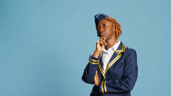 African american woman thinking about solutions, brainstorming ideas in studio. Professional airliner in uniform feeling pensive and thoughtful, pondering decision on blue backdrop.