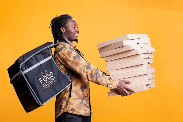 ピザ屋の宅配業者は ランチタイムに顧客のための配達を準備し ピザボックスでいっぱいのスタックを保持します 持ち帰り用の熱バックパックを持つアフリカ系アメリカ人の労働者は テイクアウト食品の注文を提供 — ストック写真