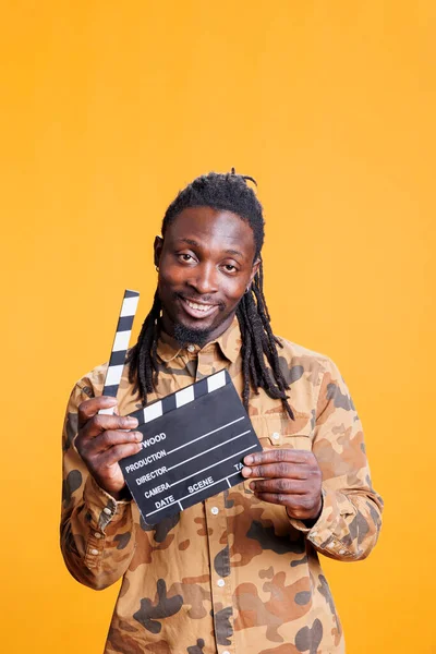 黄色の背景の上にスタジオに立って映画のクラッパーボードを保持アフリカ系アメリカ人の男の笑顔の肖像画 若い大人がエンターテイメントビデオのオーディションを受ける 映画的概念 — ストック写真