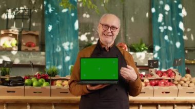 Yerel gıda fuarında, güneşli bir günde, izole edilmiş yeşil ekranlı dizüstü bilgisayarı olan yaşlı bir çiftçi. Üst düzey adam, ekranda boş kopya spreyle krom anahtar görüntüsü gösteriyor, pron giyiyor.