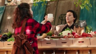 Stall holder drinking cup of hot coffee with regular customer, feeling happy with organic eco produce. Business owner sharing drink with client, selling homegrown products. Handheld shot.