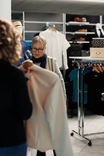 ファッショナブルな服を買いたいシニアクライアントは ショッピングセンターでカジュアルな服装で店の労働者に助けを求める 現代のブティックで新しいコレクションや流行のアクセサリーを購入する高齢女性 — ストック写真