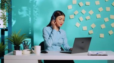 Mutlu Asyalı kadın kulaklıkla müzik dinlemekten hoşlanıyor renkli yaratıcı ofis masasında şarkı söyleyip dans ediyor dizüstü bilgisayarla çalışırken. Mavi stüdyo arka planında rahat iş yerinde çalışan iş kadını.