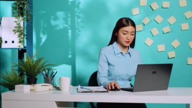 Pano veri notlarını çapraz kontrol ettikten sonra dizüstü bilgisayarda veri yazan gülümseyen bir iş kadını. Modern yaratıcı ofis masasında çalışan odaklanmış profesyonel bir çalışan mavi stüdyo geçmişi üzerinde çalışıyor.
