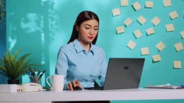 Gülümseyen profesyonel yönetici dizüstü bilgisayara veri yüklüyor, modern renkli ofis masasında kahve içiyor mavi stüdyo arka planında. Asyalı iş kadını iş sonuçlarından memnun