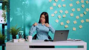 Mutlu Asyalı ofis çalışanı dövüşüyor gibi yaparak, yumruk atarak eğleniyor. Neşeli, neşeli iş kadını, mavi stüdyo arka planında rahat renkli iş yerinde boksu taklit ediyor.