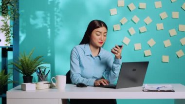 İş kadını kart bilgilerini ekliyor, çalışırken laptopta ödeme metodunu yönetiyor. Asyalı çalışan, renkli, parlak, modern ofis masasında mavi stüdyo arka planında oturuyor.