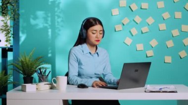 İnternetteki video görüşmesi sırasında müdürden gelen talimatları takip eden kulaklık takan bir sekreter. Asyalı bir çalışan, mavi stüdyo geçmişi üzerine renkli ofisinde patronuyla görüntülü konferans yapıyor.