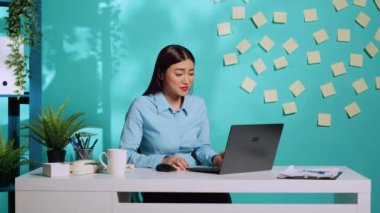 Çevrimiçi video görüşmesinde neşeli bir ofis çalışanı iş arkadaşıyla rahatlatıcı bir konuşma yapıyor. Gülümseyen bir çalışan mavi stüdyo arka planında modern işyerindeki masasında oturuyor.